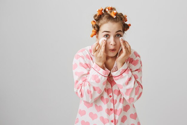 Nettes glückliches Mädchen in Nachtwäsche und Lockenwicklern wischen Make-up mit Wattepads ab