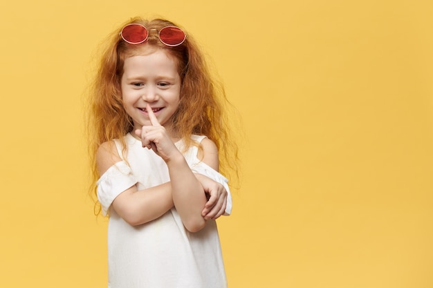 Nettes hübsches verspieltes kleines Mädchen mit stilvoller Sonnenbrille auf ihrem Kopf, das Zeigefinger an ihrem Mund hält und Stille Geste macht