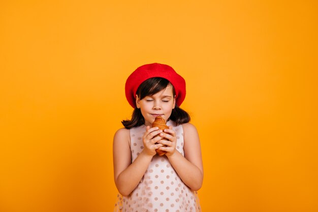 Nettes hungriges Kind, das Croissant isst. dunkelhaariges kleines Mädchen isoliert auf gelber Wand.