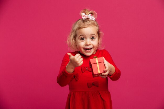 Nettes junges blondes Mädchen im roten Kleid, das Geschenkbox hält