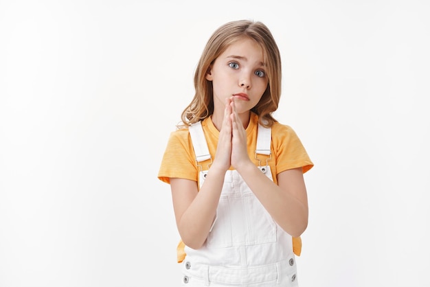 Nettes kleines anhängliches Mädchen, junge blonde Tochter hält Hände, betet, Handflächen zusammen, zieht trauriges Mitleidsgesicht, tief bittender Blick mit blauen Augen, Entschuldigung