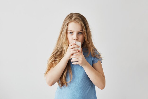 Nettes kleines blondes Mädchen mit bezaubernden blauen Augen konzentriertes Schauen und Trinken von Glas Milch. Glückliche Kindheitsmomente.