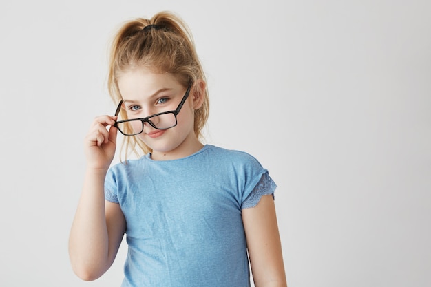 Nettes kleines blondes Mädchen mit blauen Augen und angenehmem Lächeln im blauen T-Shirt lustiges Aufstellen mit neuer Brille für Schulfoto.