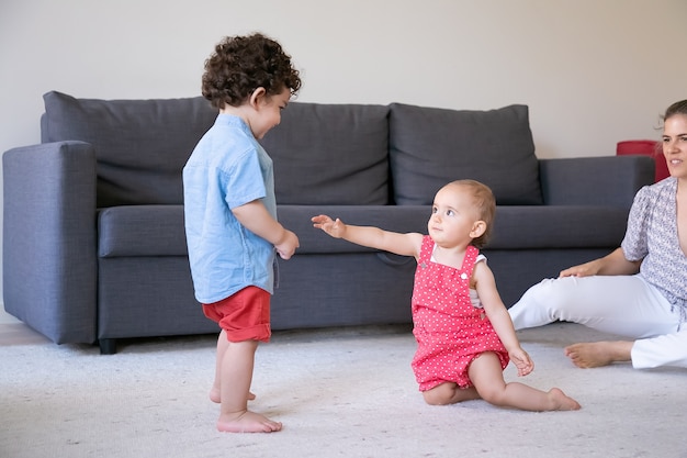 Nettes kleines Mädchen, das auf Teppich mit gemischtem Jungen spielt. Beschnittene junge Mutter, die Kinder beobachtet und lächelt. Lockiges Kind, das barfuß im Wohnzimmer steht. Familienhaus, Wochenend- und Kindheitskonzept