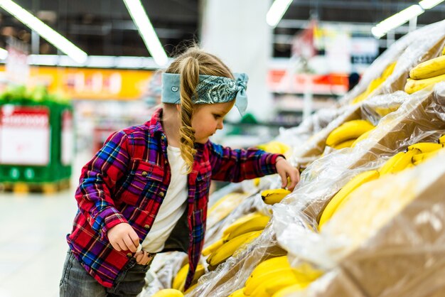 Nettes kleines Mädchen, das Bananen in einem Lebensmittelgeschäft oder im Supermarkt hält