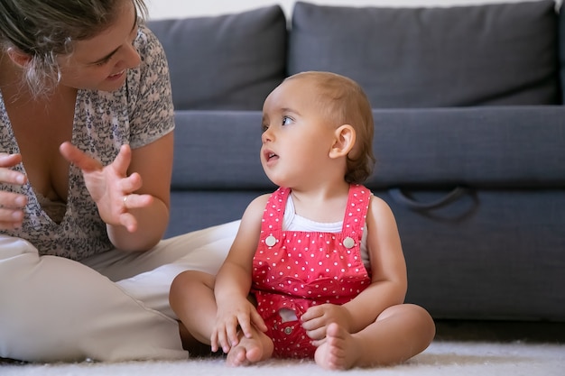 Nettes kleines Mädchen, das Mutter mit offenem Mund zuhört und sie ansieht. Beschnittene Mutter, die mit gekreuzten Beinen auf dem Boden sitzt und mit der Tochter spricht. Schönes Kind, das barfuß sitzt. Wochenend- und Mutterschaftskonzept