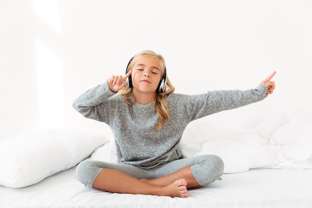 Nettes kleines Mädchen im grauen Pyjama, das Musik mit geschlossenen Augen hört, während in ihrem Bett sitzend