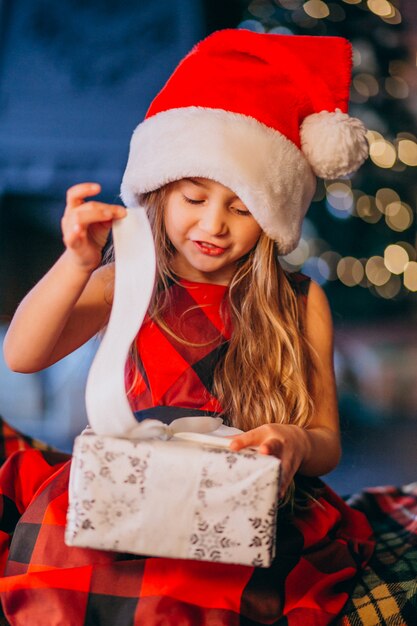 Nettes kleines Mädchen im Sankt-Hut, der Weihnachtsgeschenk auspackt