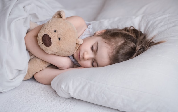 Nettes kleines Mädchen schläft süß in einem weißen gemütlichen Bett mit einem weichen Bärenspielzeug, dem Konzept der Kinderruhe und des Schlafes