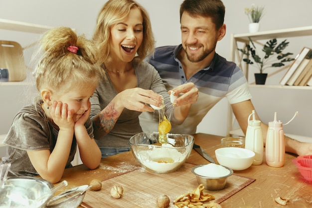 Nettes kleines Mädchen und ihre schönen Eltern bereiten den Teig für den Kuchen in der Küche zu Hause vor. Familienlebensstilkonzept