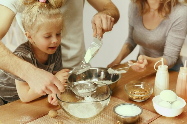 Nettes kleines Mädchen und ihre schönen Eltern bereiten den Teig für den Kuchen in der Küche zu Hause vor. Familienlebensstilkonzept