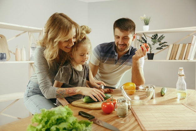 Nettes kleines Mädchen und ihre schönen Eltern schneiden Gemüse und lächeln, während sie Salat in der Küche zu Hause machen. Familienlebensstilkonzept