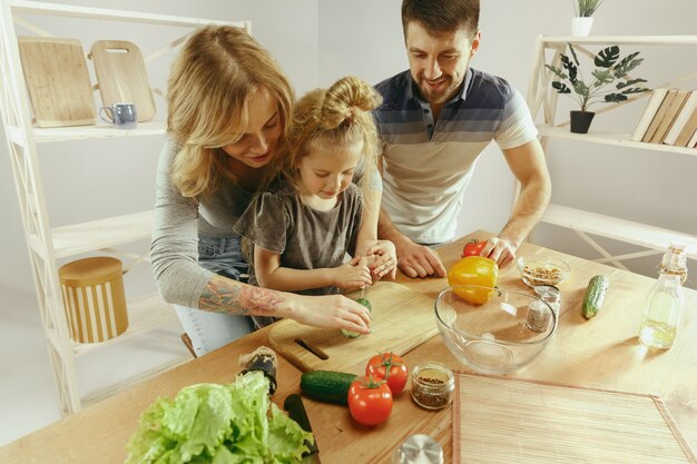 Nettes kleines Mädchen und ihre schönen Eltern schneiden Gemüse und lächeln, während sie Salat in der Küche zu Hause machen. Familienlebensstilkonzept