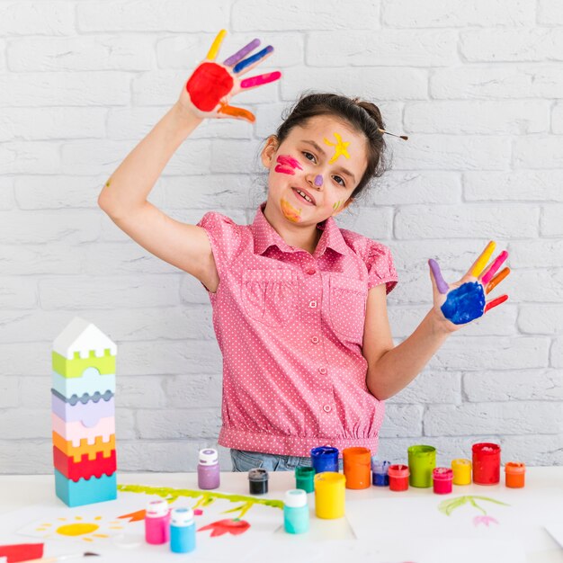 Nettes kleines Mädchen, welches die gemalten Hände steht vor Tabelle mit bunten Farben zeigt