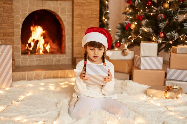 Nettes kleines weibliches Kind mit weißem Pullover und Weihnachtsmann-Hut, das im festlichen Raum mit Kamin und Weihnachtsbaum posiert, Geschenkbox in den Händen hält und mit Erstaunen in die Gegenwart schaut.