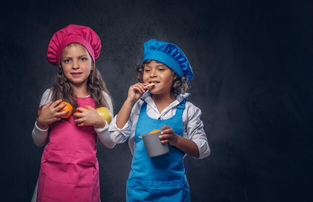 Nettes Kochpaar. Kleiner Junge mit braunem, lockigem Haar, gekleidet in eine blaue Kochuniform und ein schönes Schulmädchen, gekleidet in eine rosafarbene Kochuniform, posiert mit Kochgeschirr und Früchten in einem Studio. Isoliert auf einem dar