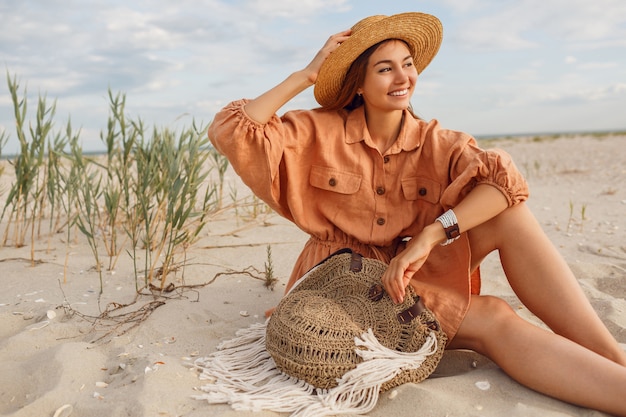 Nettes lächelndes Mädchen im stilvollen Leinenkleid, das am Abendstrand auf weißem Sand entspannt. Strohhut, Boho-Tasche. Feiertage mod.