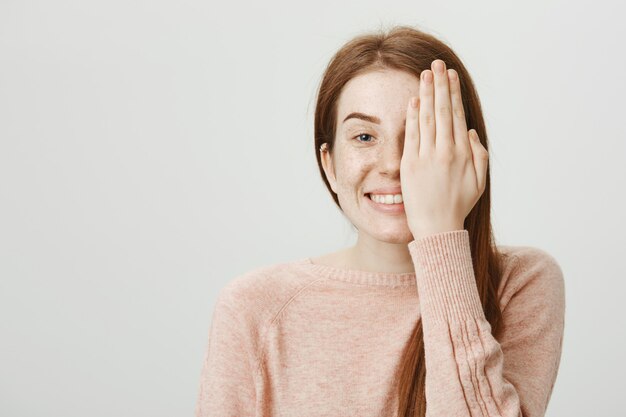 Nettes lächelndes rothaariges Mädchen, das ihre Sicht am Optikerladen überprüft, bedeckt Gesichtshälfte mit der Hand