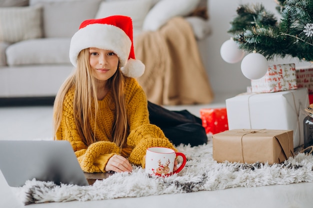 Nettes Mädchen, das Computer durch Weihnachtsbaum benutzt