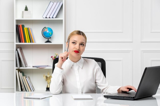 Nettes Mädchen des Kundendienstes im weißen Hemd mit rotem Lippenstift und Laptop, der mit Stift auf Schläfe denkt