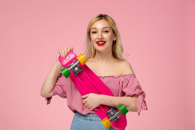 Nettes Mädchen entzückendes blondes Küken mit rotem Lippenstift in der rosa Bluse, die glücklich rosa Skateboard hält