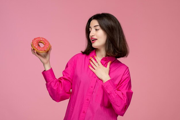 Nettes Mädchen hübsches entzückendes Mädchen im rosa Hemd mit rotem Lippenstift, der Donut liebevoll betrachtet