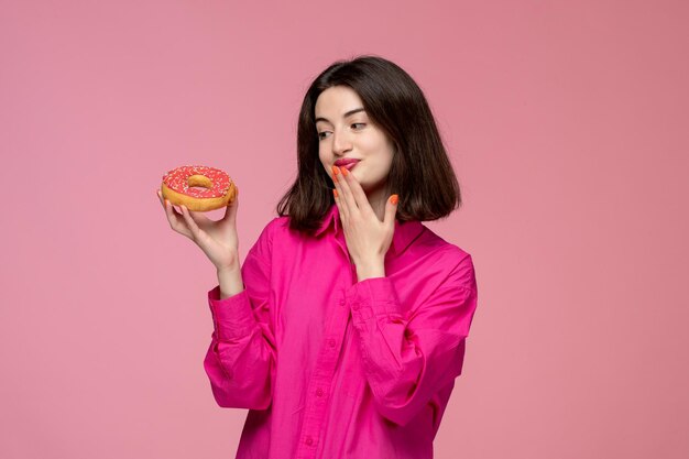 Nettes Mädchen hübsches junges schönes Brunettemädchen im rosafarbenen Hemd, das daran denkt, einen Donut zu essen