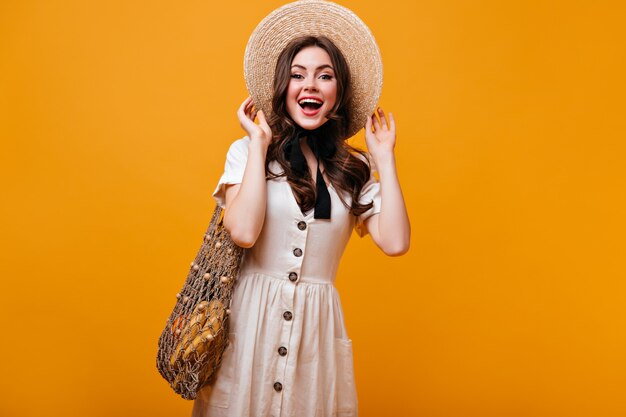 Nettes Mädchen im Baumwollkleid, das mit Stringtasche mit Früchten aufwirft. Frau im Strohhut mit Bogen lacht auf orange Hintergrund.