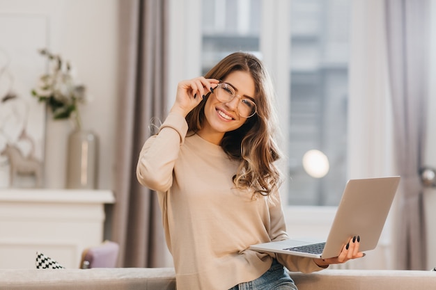 Nettes Mädchen im beige Hemd, das Gläser berührt und Laptop mit Lächeln hält