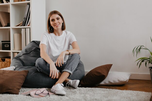 Nettes Mädchen im hellen Straßenoutfit, entspannt im grauen Stuhl auf Regal mit Ordnern und Büchern sitzend.