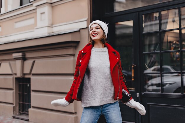 Nettes Mädchen in Jeans, grauem Pullover, rotem Mantel und Strickmütze mit Handschuhen, die mit Lächeln auf der Straße aufwerfen.