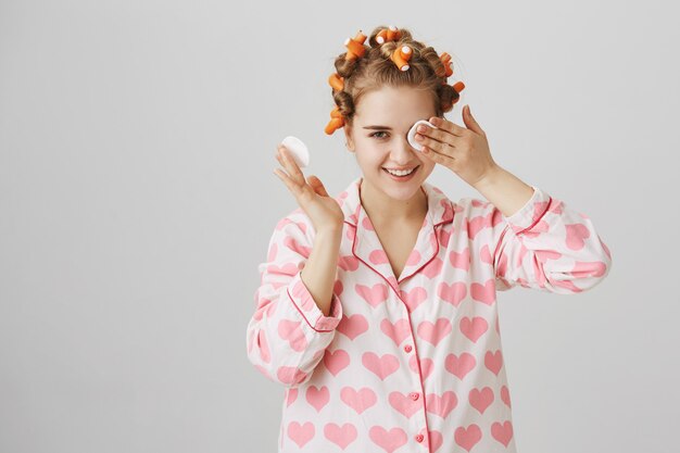 Nettes Mädchen-Make-up vor dem Schlafengehen mit Wattepad, Lockenwickler und Pyjama tragend