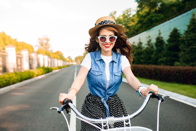 Kostenloses Foto nettes mädchen mit dem langen lockigen haar in der sonnenbrille, die ein fahrrad zur kamera auf straße fährt. sie trägt einen langen rock, ein wams und einen hut. sie sieht glücklich aus.