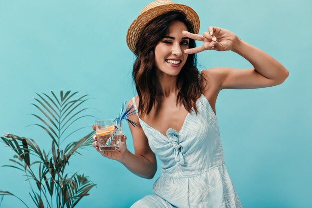 Nettes Mädchen zeigt Friedenszeichen, zwinkert und hält Wasser mit Orange. Lächelnde Frau mit dem kurzen dunklen Haar, das nahe kleiner Palme aufwirft.