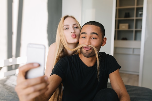 Nettes Paar, das Selfie am Telefon auf Bett in der modernen Wohnung macht. Hübscher Kerl, der Spaß mit langen blonden Haaren von seiner Freundin hat und einen Kuss, Freude, fröhliche Stimmung sendet