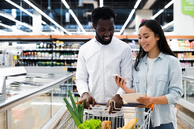 Nettes Paar mit Warenkorb überprüfend auf beweglicher Einkaufsliste am Supermarkt