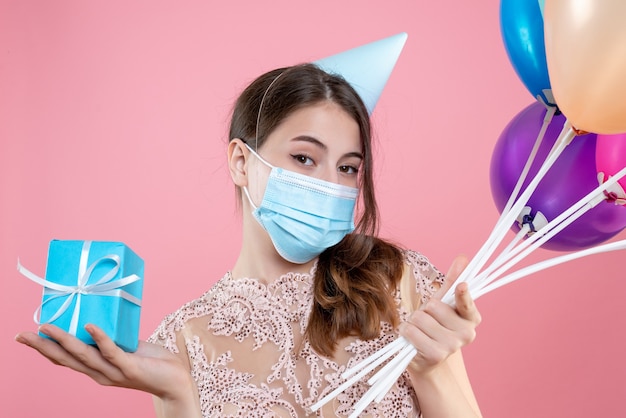 Nettes Partygirl der Nahaufnahme-Vorderansicht mit Krone und Maske, die Luftballons und Geschenk halten