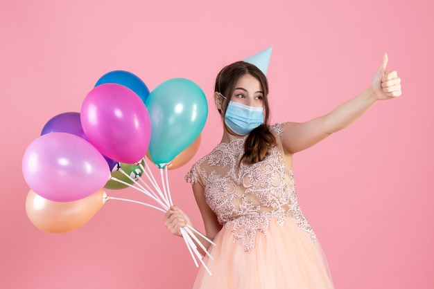 Nettes Partygirl der Vorderansicht mit der Partykappe, die bunte Luftballons hält, die Daumen hoch Zeichen machen