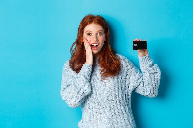 Nettes rothaariges Mädchen im Pullover, der Kreditkarte zeigt, in die Kamera lächelt und auf blauem Hintergrund steht