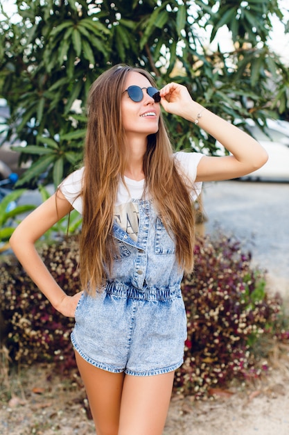 Nettes schlankes gebräuntes Mädchen, das nahe Baum steht. Sie trägt ein weißes T-Shirt, Jeansshorts und eine schwarze Sonnenbrille