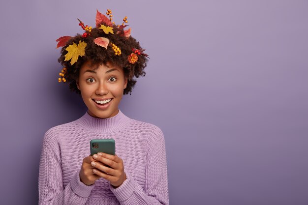 Nettes schönes dunkelhäutiges Mädchen hält modernes Handy, schaut direkt in die Kamera, wird telefonieren, lächelt glücklich, in lässigem Outfit gekleidet, posiert während der Herbstzeit, isoliert auf lila Wand