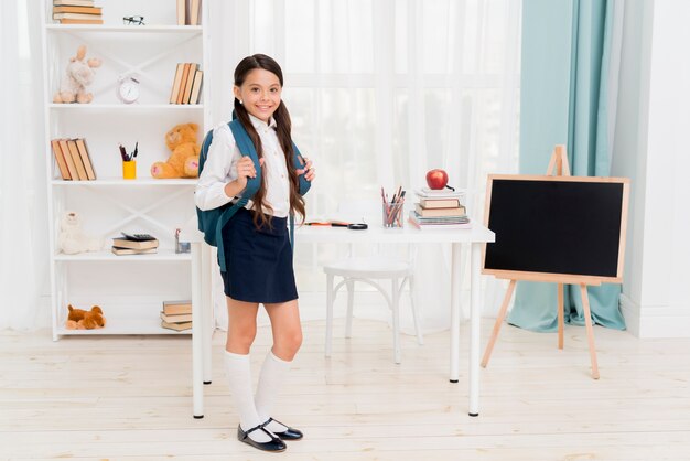 Nettes Schulkind mit dem Rucksack, der vor Schreibtisch am Klassenzimmer steht