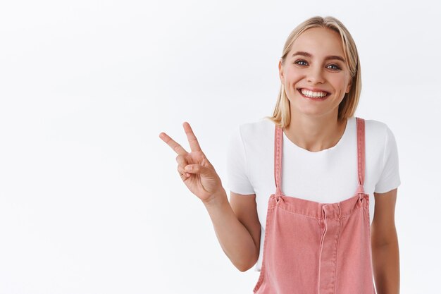 Nettes und reizendes blondes kaukasisches Mädchen in rosa Latzhose, T-Shirt, das schüchtern und zärtlich lächelt, Sieg- oder Friedenszeichen zeigt, Goodwill-Bewegung, posiert für Foto, das auf weißem Hintergrund steht