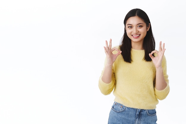 Nettes und zartes koreanisches Mädchen stimme dir zu, gib die Erlaubnis. Schöne asiatische Frau, die okaygesten zeigt und lächelt, Wahl genehmigen, Produkt empfehlen, positive Bewertung hinterlassen, weiße Wand