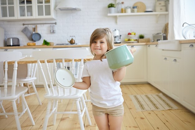 Nettes weibliches Kind in der Freizeitkleidung, die Auflauf und Halter hält, aufgeregten Blick hat, Suppe kochend, moderne Küche