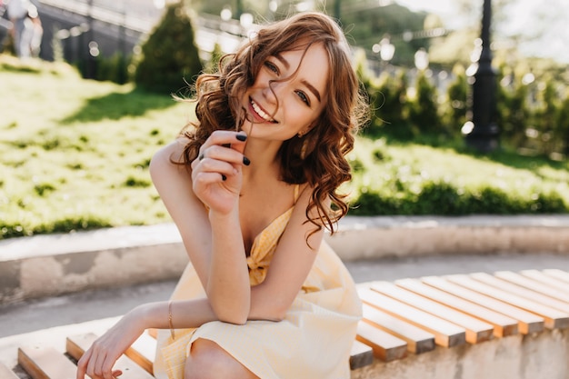 Nettes weibliches Modell mit gewelltem rotem Haar, das auf Parkbank sitzt. Foto im Freien des glückseligen Ingwermädchens im gelben Kleid, das auf Natur aufwirft.