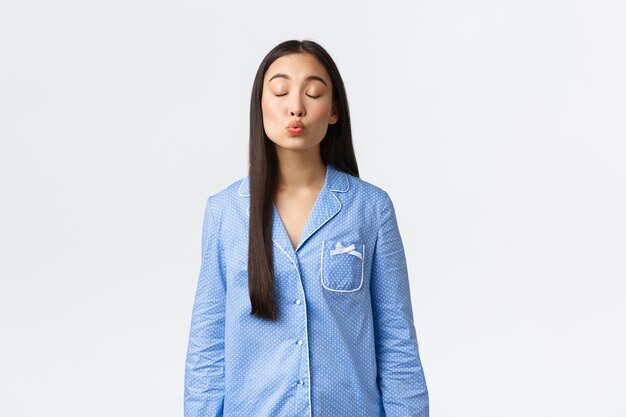 Nettes zartes asiatisches mädchen in blauen pyjamas, das nachts einen romantischen traum hat, augen schließt und jemanden küsst, freund oder datum abbildet, weißer hintergrund steht, tagträumt.