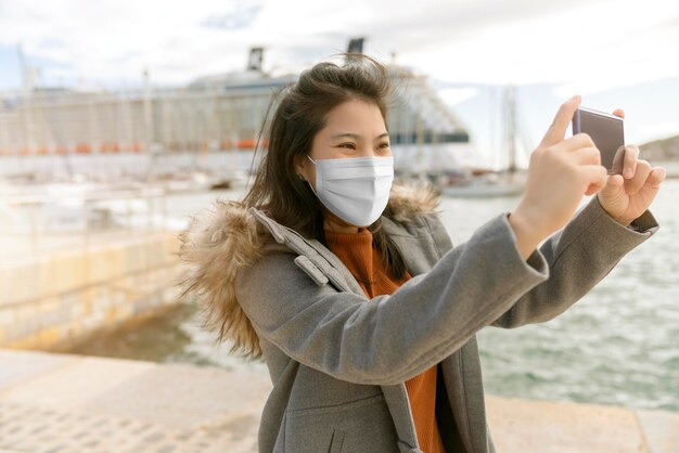 Neue asiatische frau des normalen reiselebensstils mit maske covid19 schutzhandgriff smartphonekamera oder kaffeetassenreisebesichtigung mit hintergrund des kreuzfahrtschiffes am seehafen