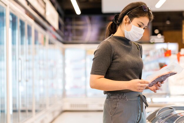 Neue Normalität nach Covid-Epidemie Junge intelligente asiatische Frau kauft neuen Lebensstil im Supermarkt mit Gesichtsschild oder Maskenschutz Hand wählt frisches Obstgemüse neuen normalen Lebensstil