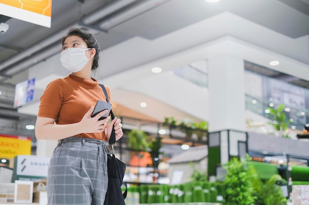 Neuer normaler Lebensstil asiatisches weibliches braunes Kleid trägt Schutzmaske beim Einkaufen im Kaufhaus nach der Quarantänezeit der Sperrung Neuer normaler Lebensstil Hand verwenden Smartphone Bokeh-Hintergrund